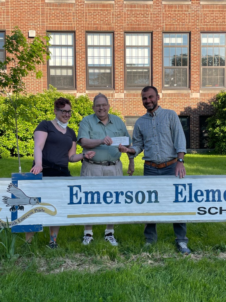 Three people stand right behind the Emerson Elementary School sign. From left to right are the neighborhood association cochair, the Giving Committee Chair, and a representative from the Emerson Elementary PTO. The school is in the background. 